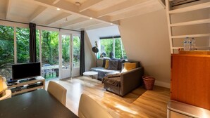Un salon moderne avec un canapé, un téléviseur et une table à manger. Une grande fenêtre offre une vue sur la verdure environnante.