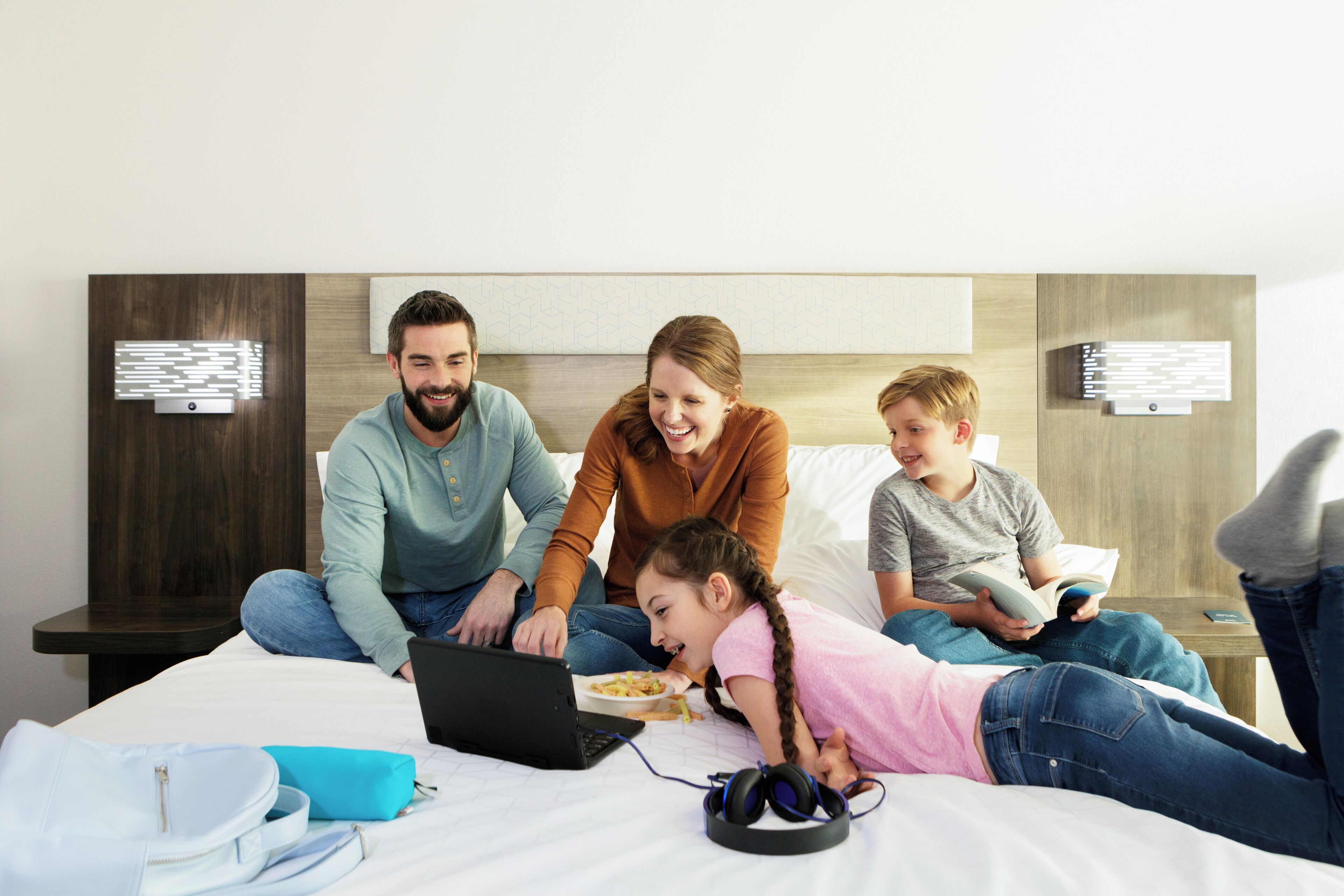 A family of four in a hotel room with a bed, a laptop, and a child playing.