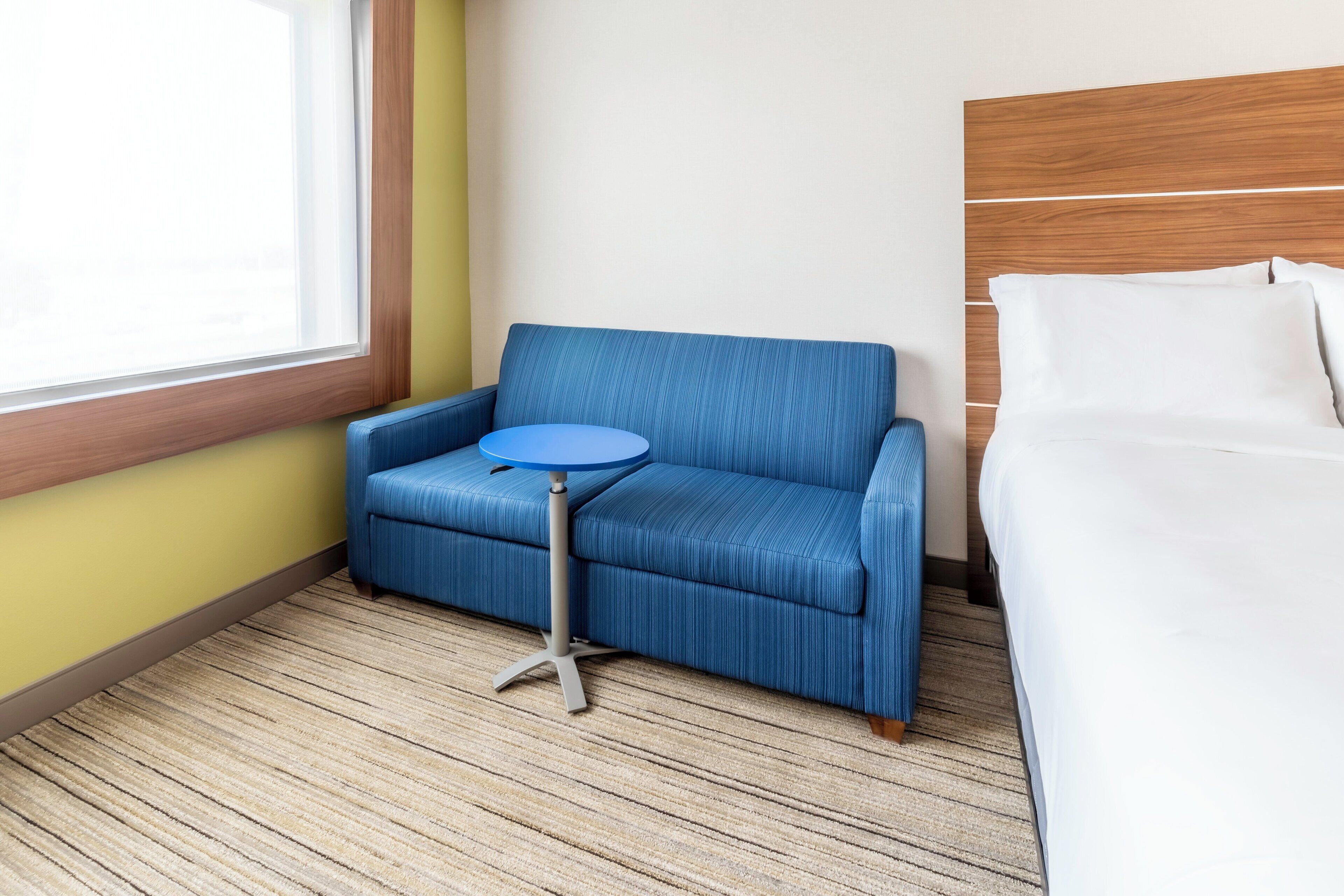 A hotel room with a blue armchair, a small round table, a bed with white bedding, and a window with blinds.
