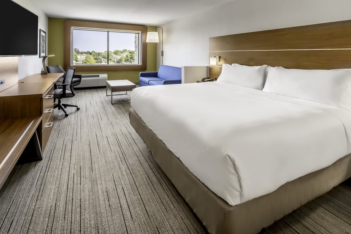 A hotel room with a large bed, a desk with a chair, a small table, and a window with a view of greenery.