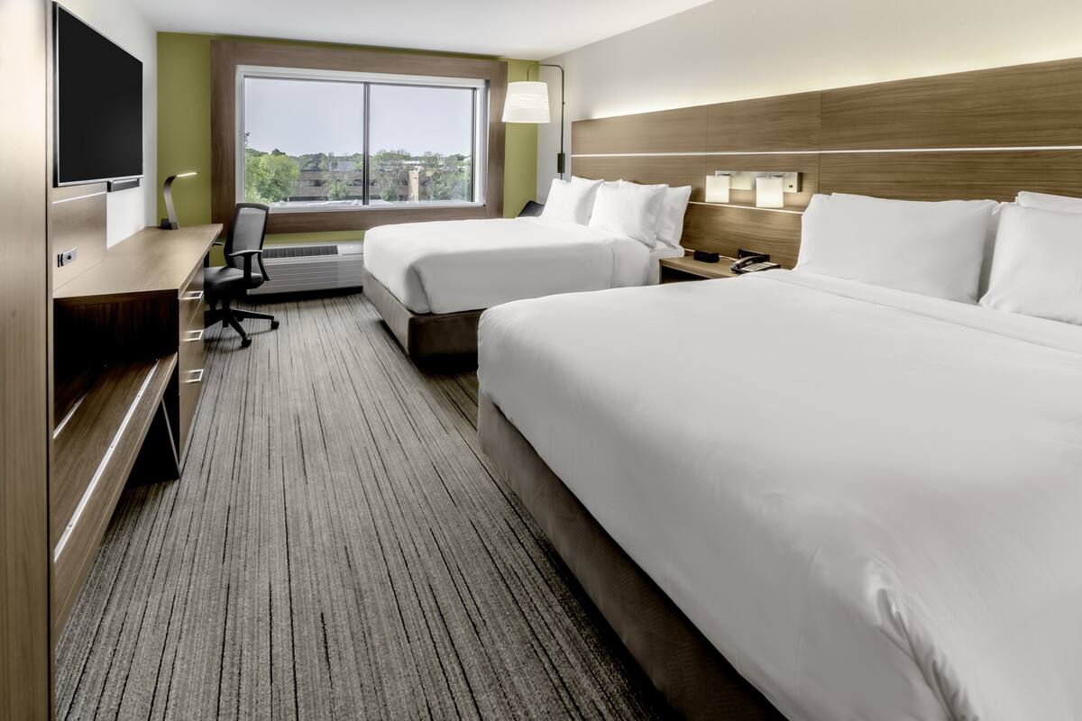 A hotel room with two beds, a desk, a chair, a TV, and a large window offering a city view.