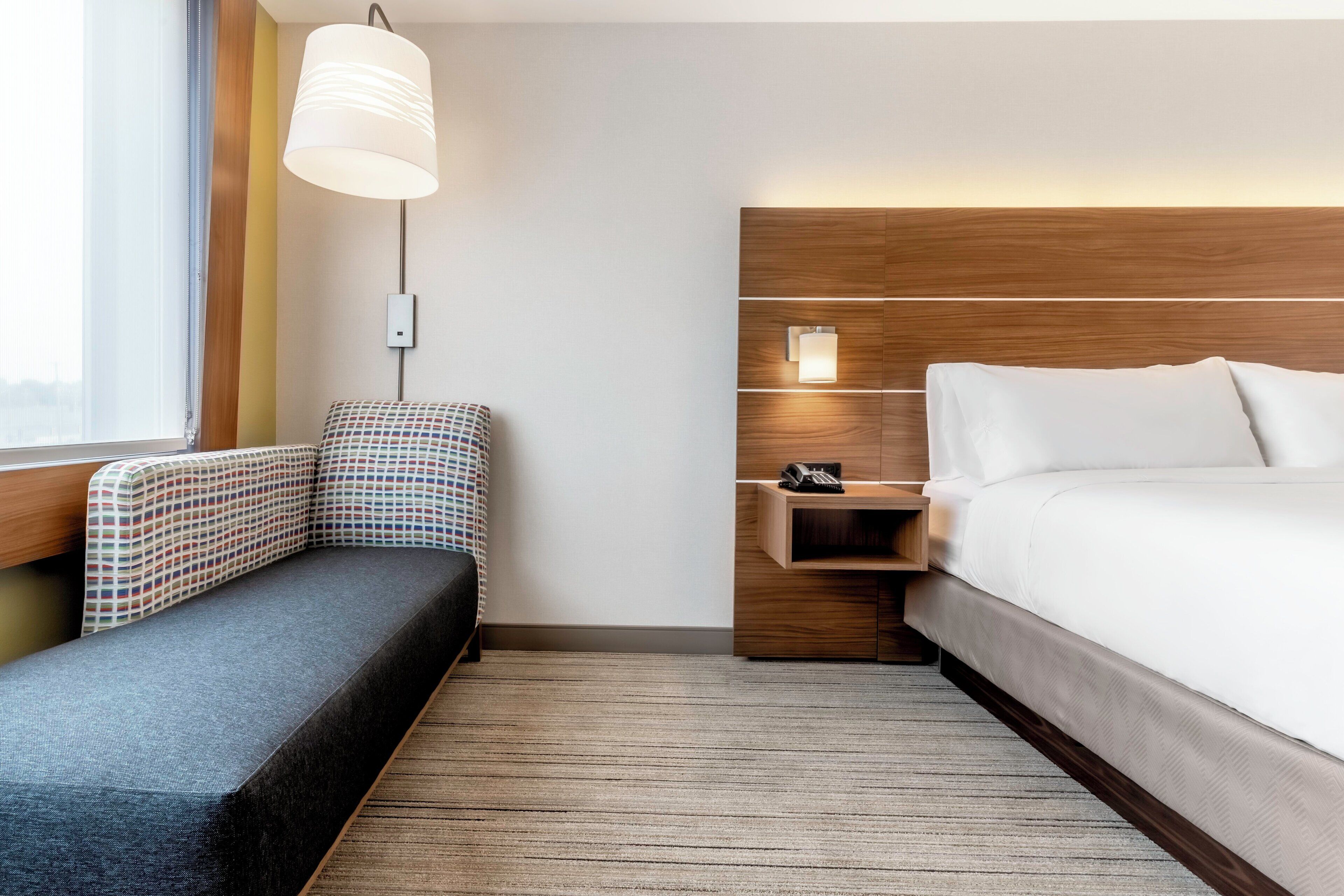 A hotel room with a bed, a desk with a telephone, a chair, a lamp, and a window with curtains.