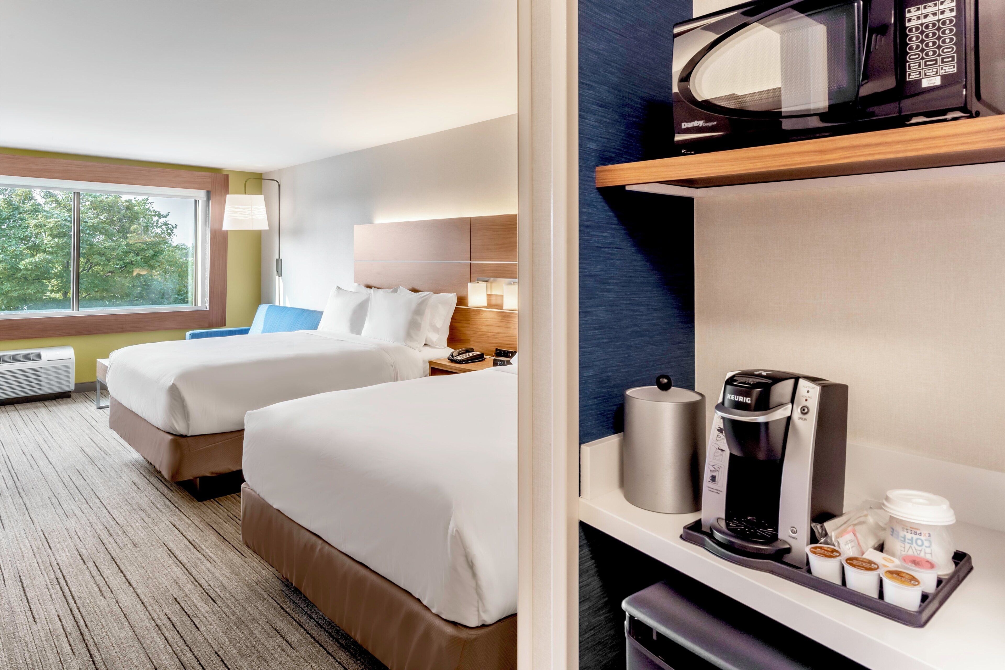 Ein Hotelzimmer mit zwei Betten, einer Mikrowelle, einer Kaffeemaschine und einem Fenster mit Blick auf Bäume.