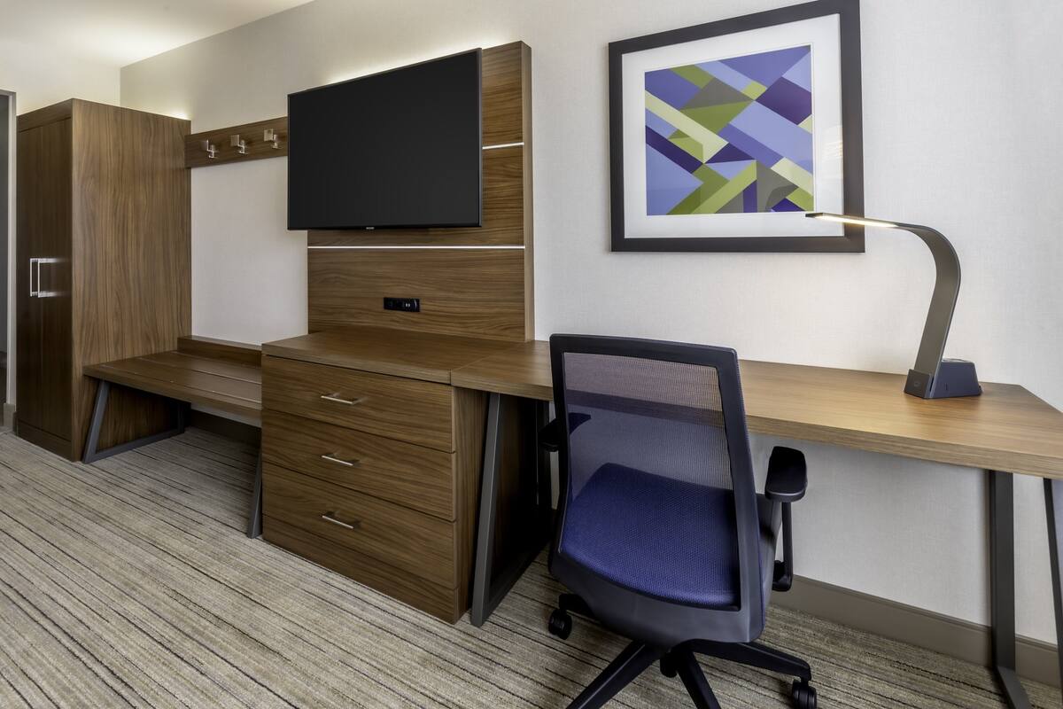 A modern office space with a wooden desk, a blue office chair, a mounted TV, and a framed abstract art piece on the wall.