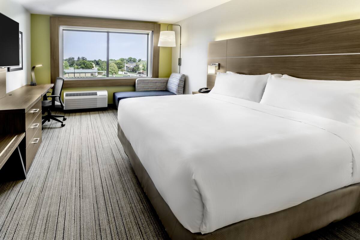 A hotel room with a large bed, a desk with a chair, a television, and a window with a view of trees and buildings.