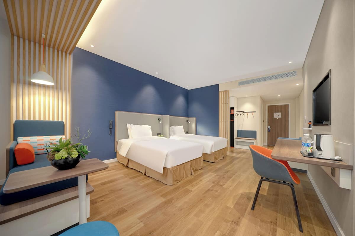 A modern hotel room with two beds, a desk, a chair, a TV, and a plant.