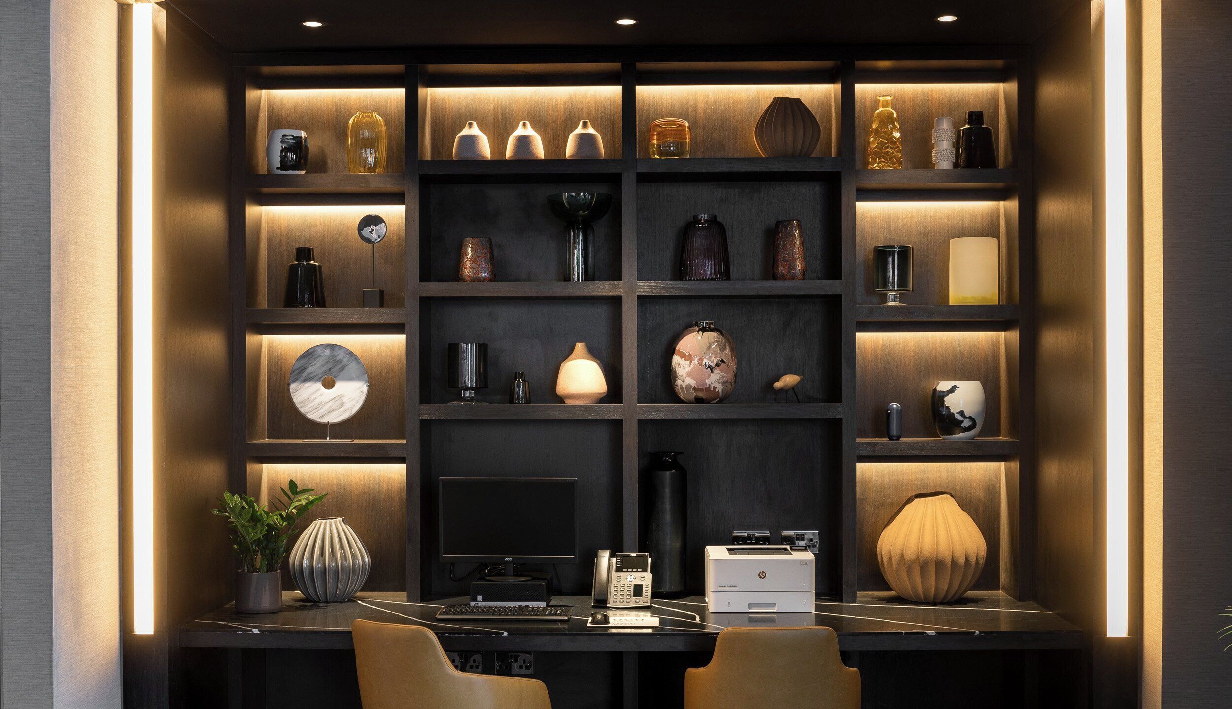 A modern office space with a desk, chairs, a computer, printer, and various decorative items on shelves.