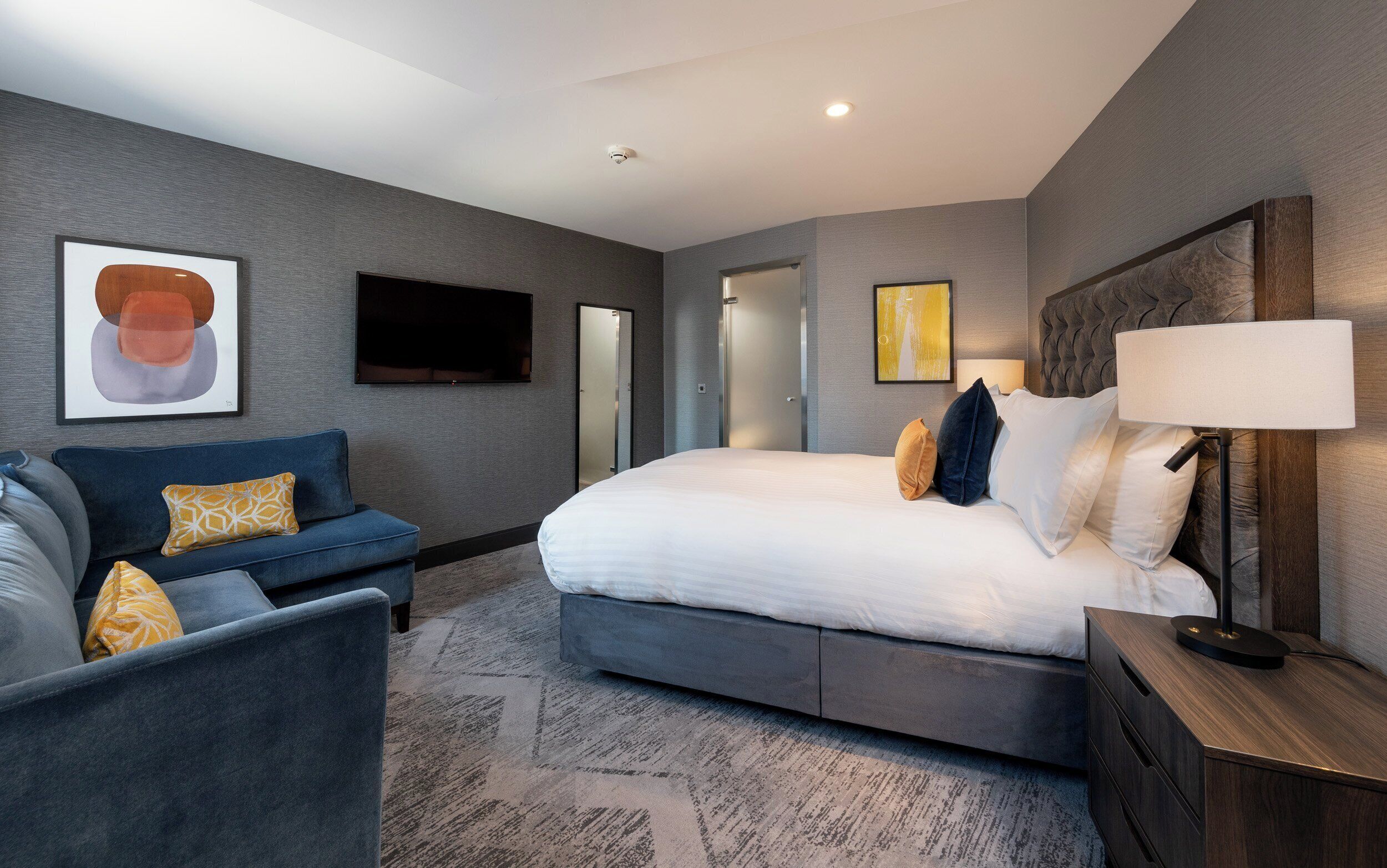 A modern hotel room with a bed, a sofa, a TV, and artwork on the walls.