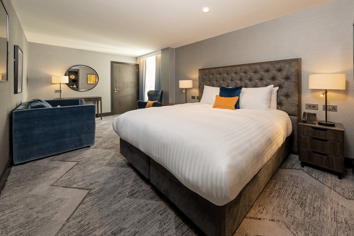 A hotel room with a large bed, a sofa, a desk, and a mirror.