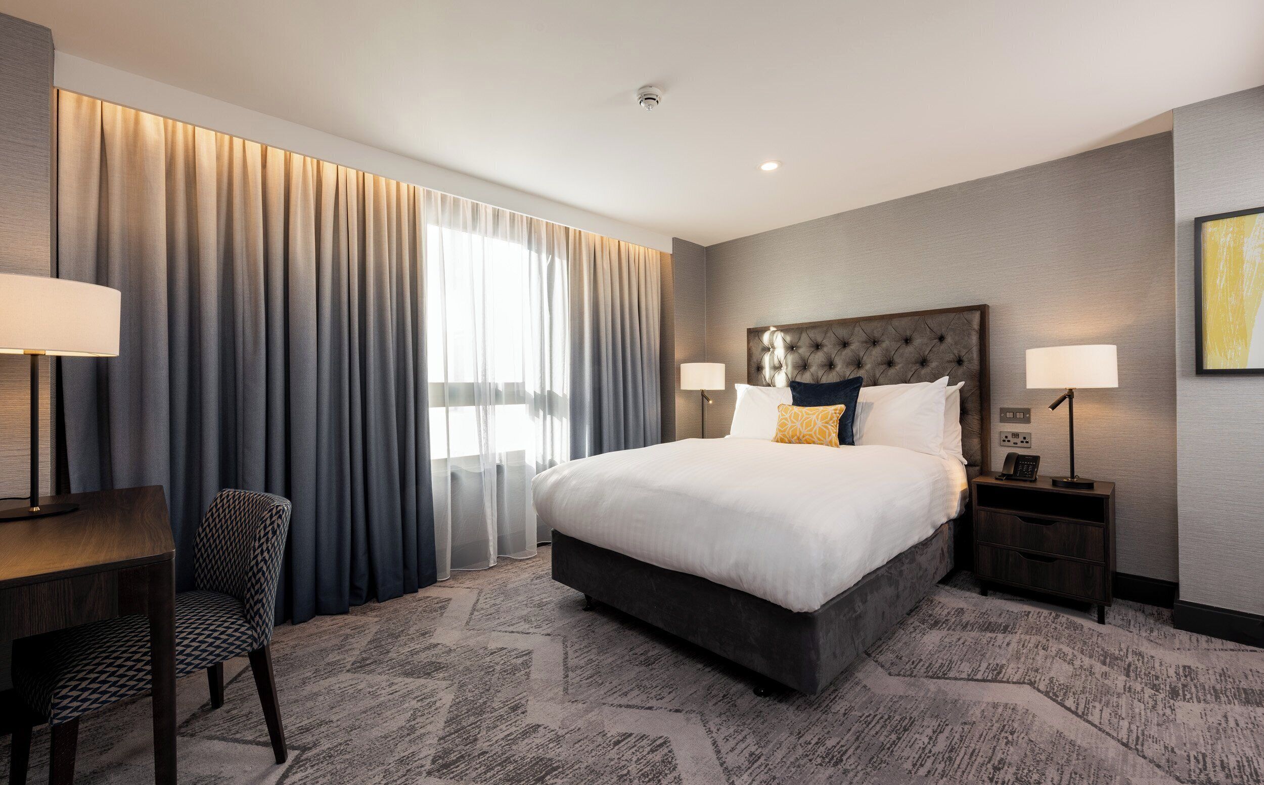 A hotel room with a large bed, a desk, a chair, and a nightstand with a lamp.