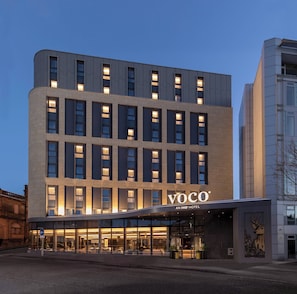 Un hôtel moderne, avec une façade sobre, de grandes fenêtres et une enseigne « Voco » bien visible.