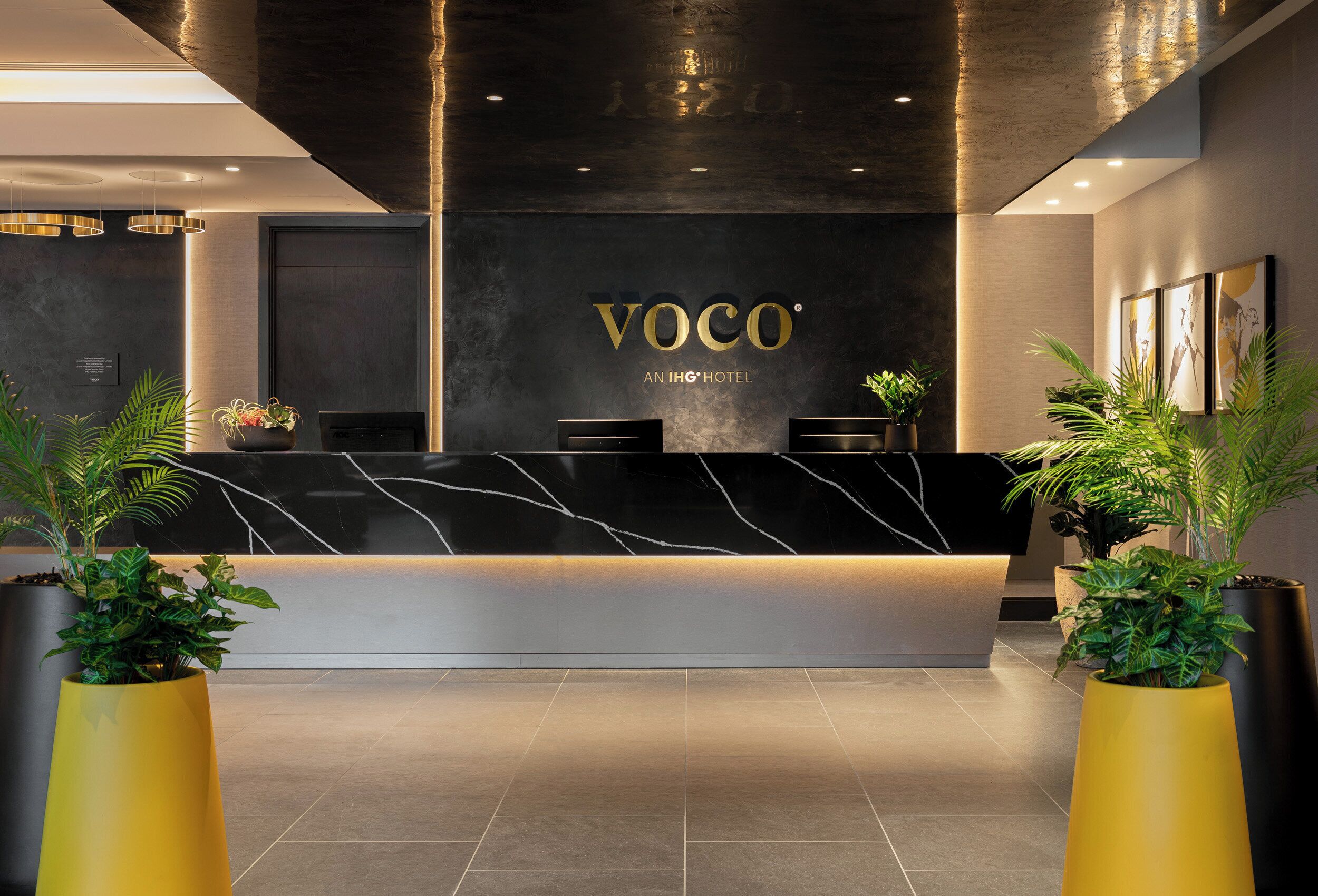 Ein moderner Hotel-Empfangsbereich mit Marmor-Theke, Zimmerpflanzen und einem Schild mit der Aufschrift 'VOCO AN IHG HOTEL'.