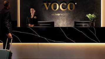 A reception desk with the 'VOCO' logo and 'AN IHG HOTEL' text, a receptionist, and a man with a suitcase.