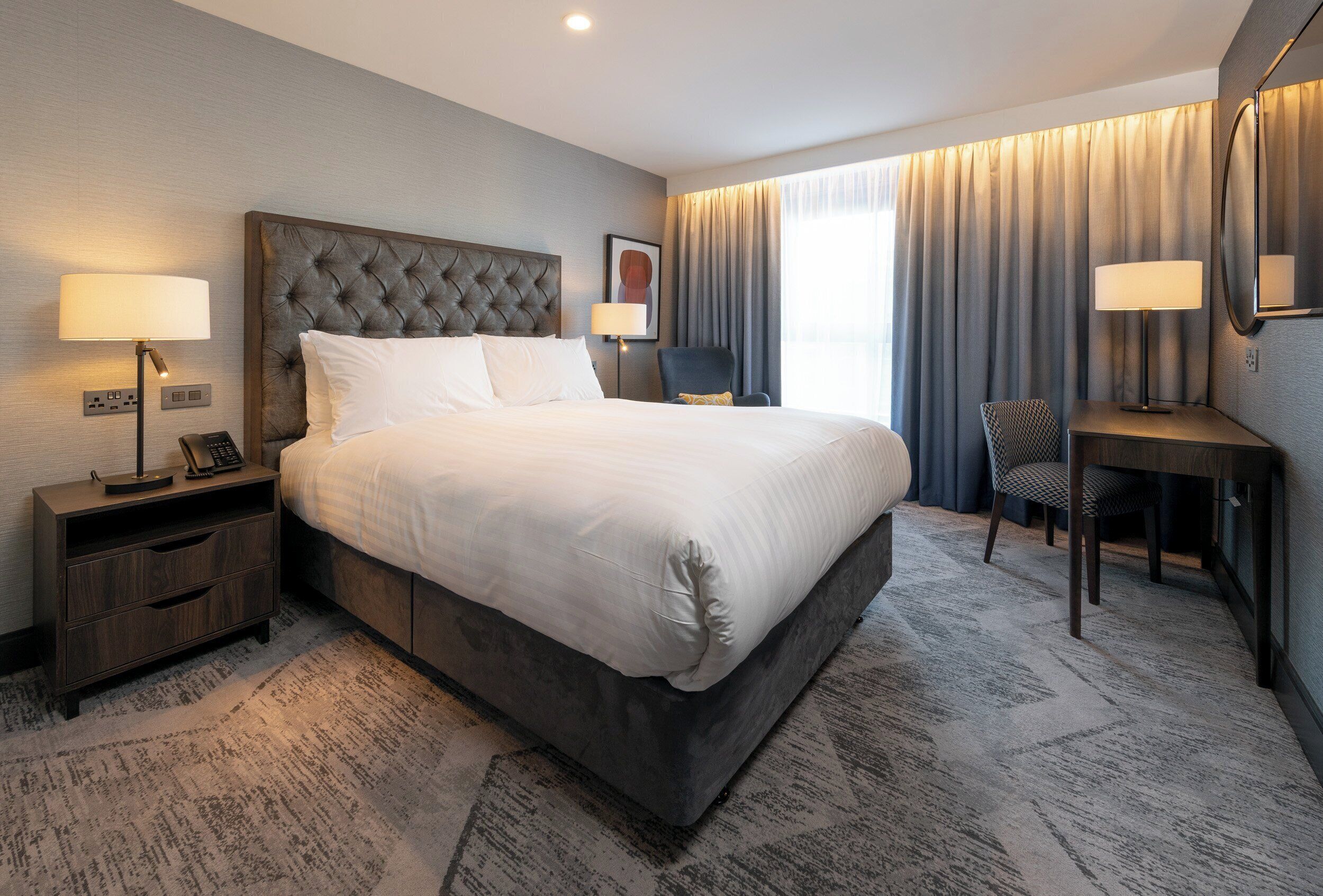 A hotel room with a large bed, bedside tables, a desk, a chair, and a window with curtains.