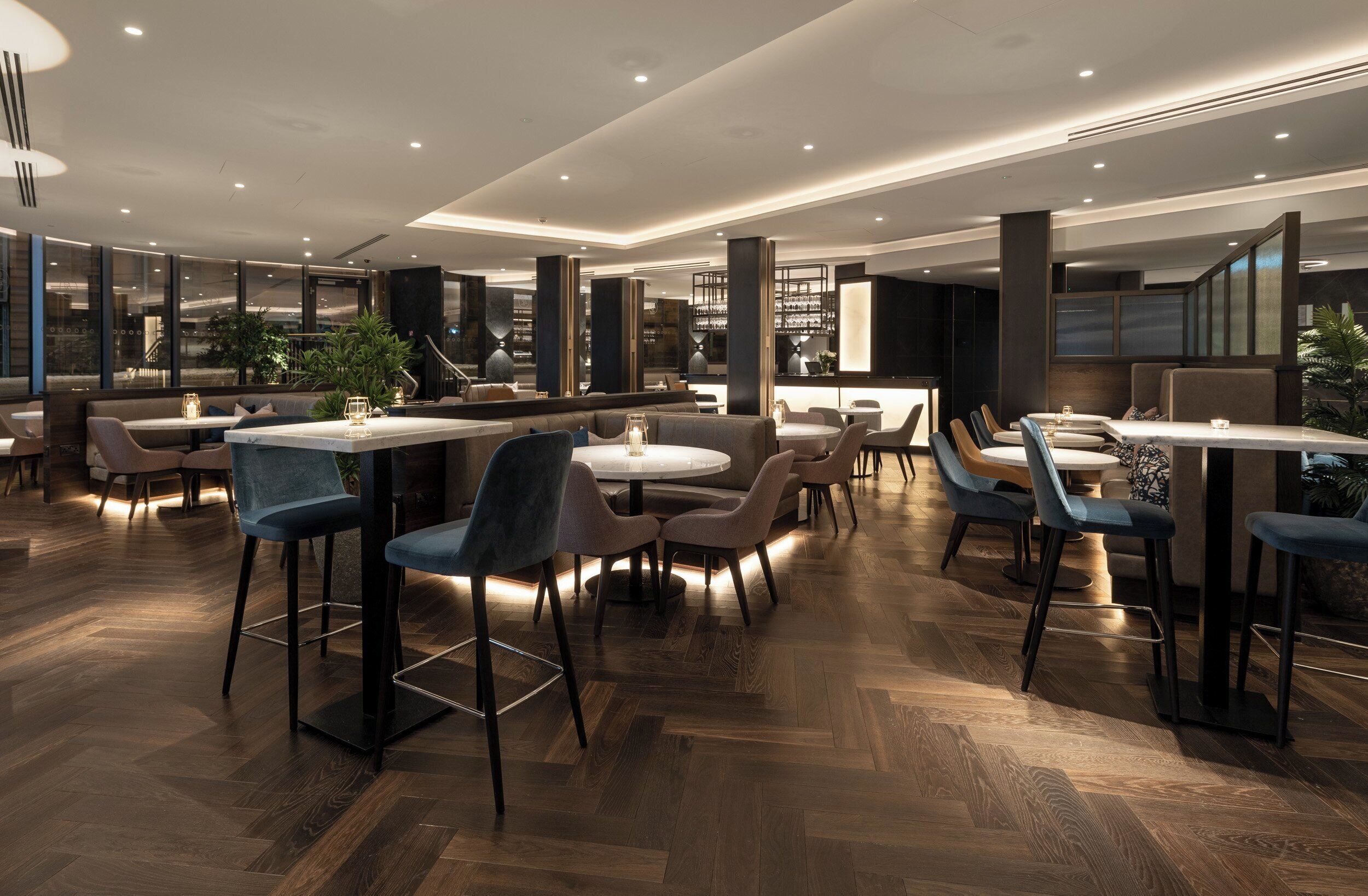 Restaurante moderno com pé direito alto, iluminação embutida e diversas opções de assentos, incluindo mesas e cabines.