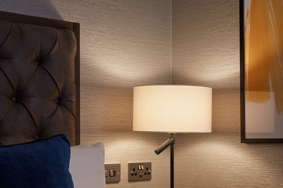 A neatly made bed with a tufted headboard, a bedside lamp, and a power outlet on the wall.