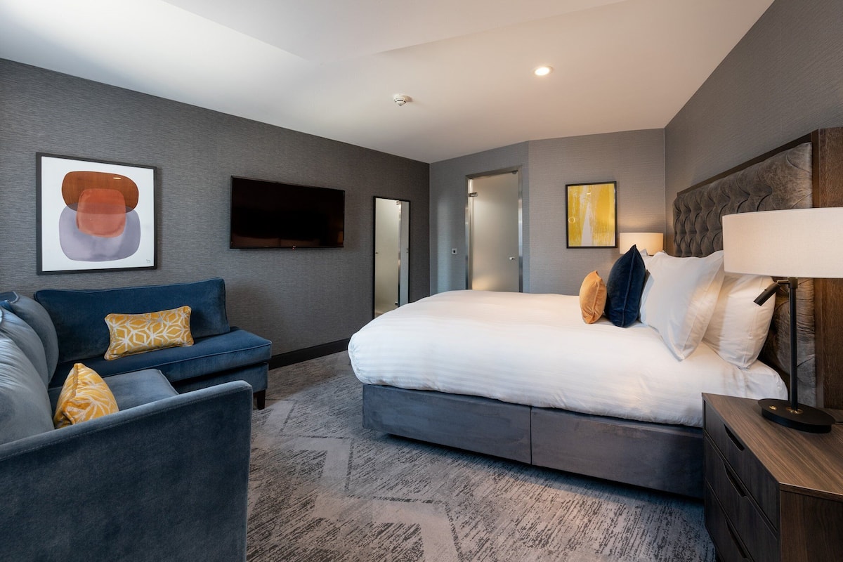 A modern hotel room with a large bed, a sofa, a TV, and a lamp.
