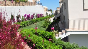 A residential building with a balcony, a garden with pink flowers, and a clear blue sky.