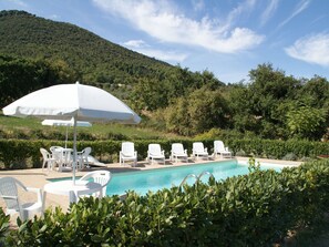 Ein Poolbereich mit weißen Liegestühlen und einem Sonnenschirm, umgeben von Grünpflanzen und einem Berg im Hintergrund.