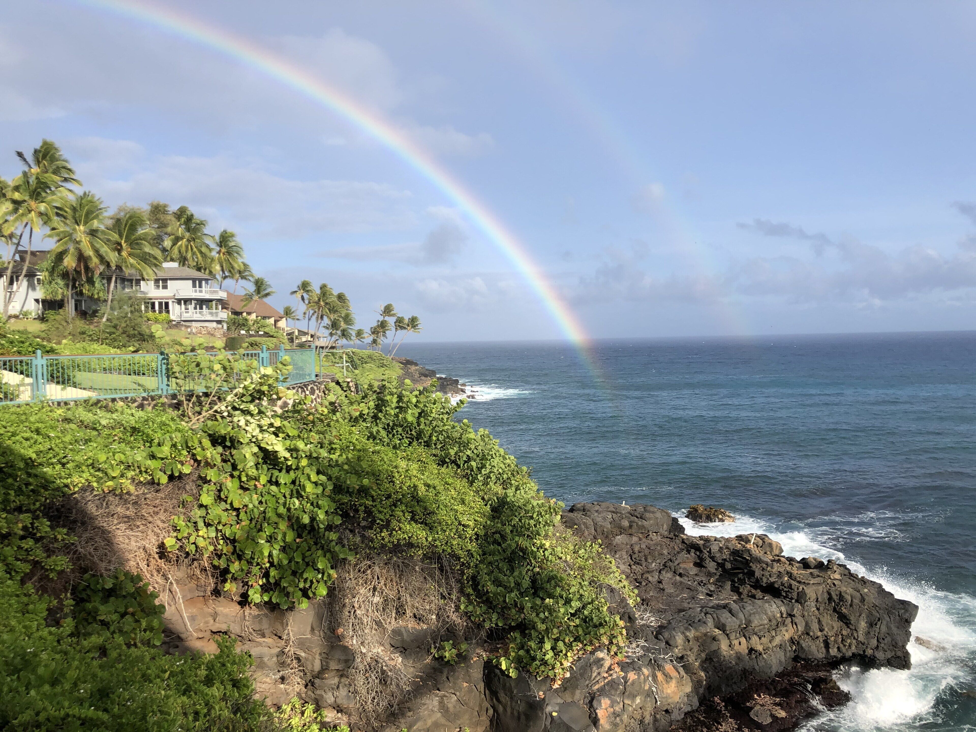 Une vue sur la côte, avec un arc-en-ciel, une végétation luxuriante et une maison offrant une vue sur l’océan.