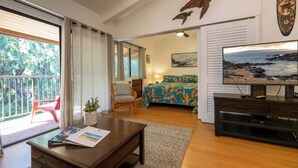 Une chambre avec un lit, une télévision, un bureau et une chaise. Il y a un balcon avec une chaise rouge et une plante.
