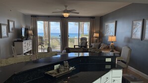 A spacious living area with a large window overlooking the ocean, a ceiling fan, and a dining area with a black countertop and white cabinets.