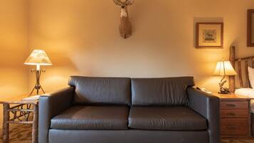 A room with a leather sofa, a mounted deer head, two lamps, a wooden nightstand, and a framed picture on the wall.