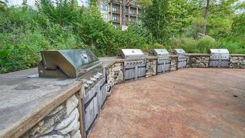A stone barbecue area with multiple grills, surrounded by greenery and a building in the background.