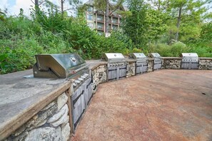A stone barbecue area with multiple grills, surrounded by greenery and a building in the background.