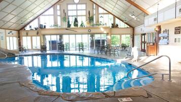 An indoor swimming pool with a high ceiling, large windows, and a seating area with tables and chairs.