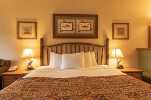 A hotel room with a wooden headboard, a patterned bedspread, two bedside lamps, and framed pictures on the walls.