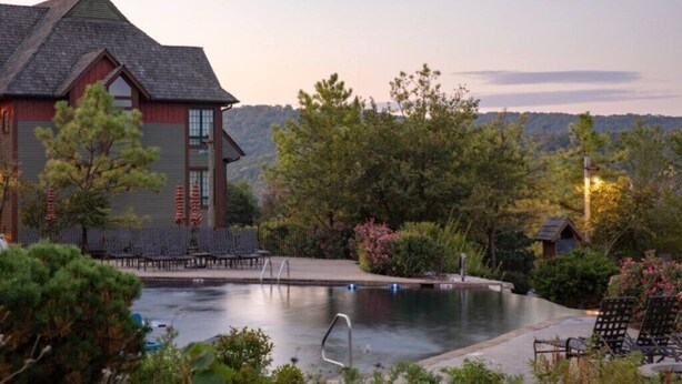 A pool area with lounge chairs, a building with a sloped roof, and surrounding trees. - Big Cedar | Rustic Retreat | Pools, Lake & Adventure (Ridgedale)