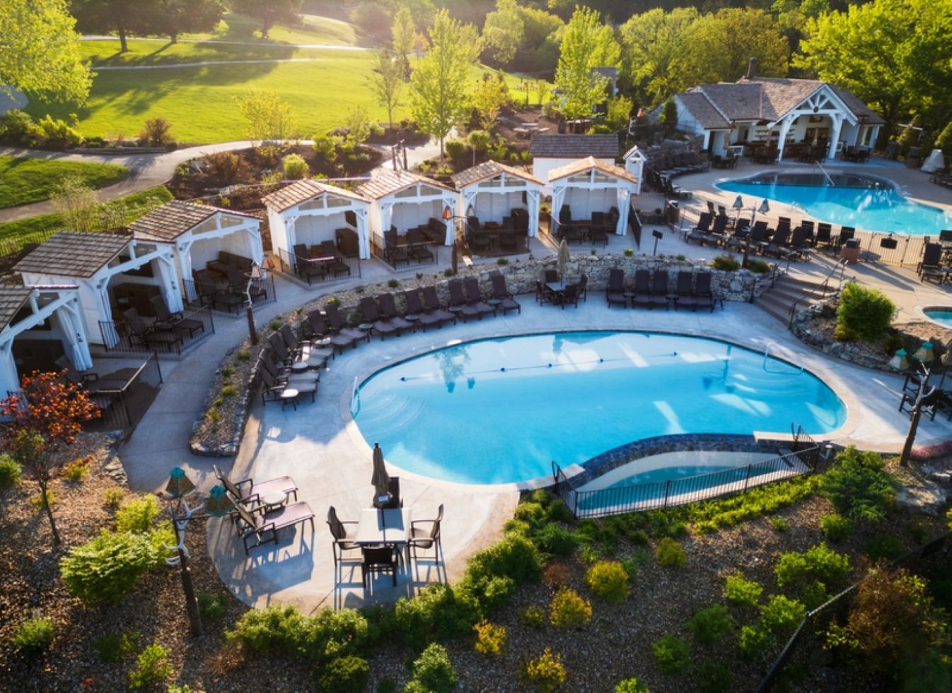 Una amplia piscina al aire libre rodeada de zonas de descanso con sillas y mesas, ubicada en un jardín ajardinado con árboles y un edificio al fondo.