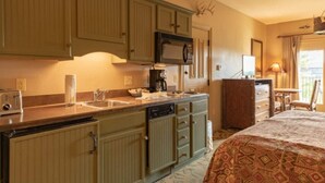 A compact hotel room with a kitchenette, a bed with patterned bedding, a desk with a lamp, and a view of the outdoors. - Big Cedar | Rustic Retreat | Pools, Lake & Adventure (Ridgedale)