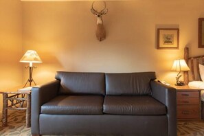 A room with a brown leather sofa, a lamp, a wooden table, and a mounted deer head on the wall.