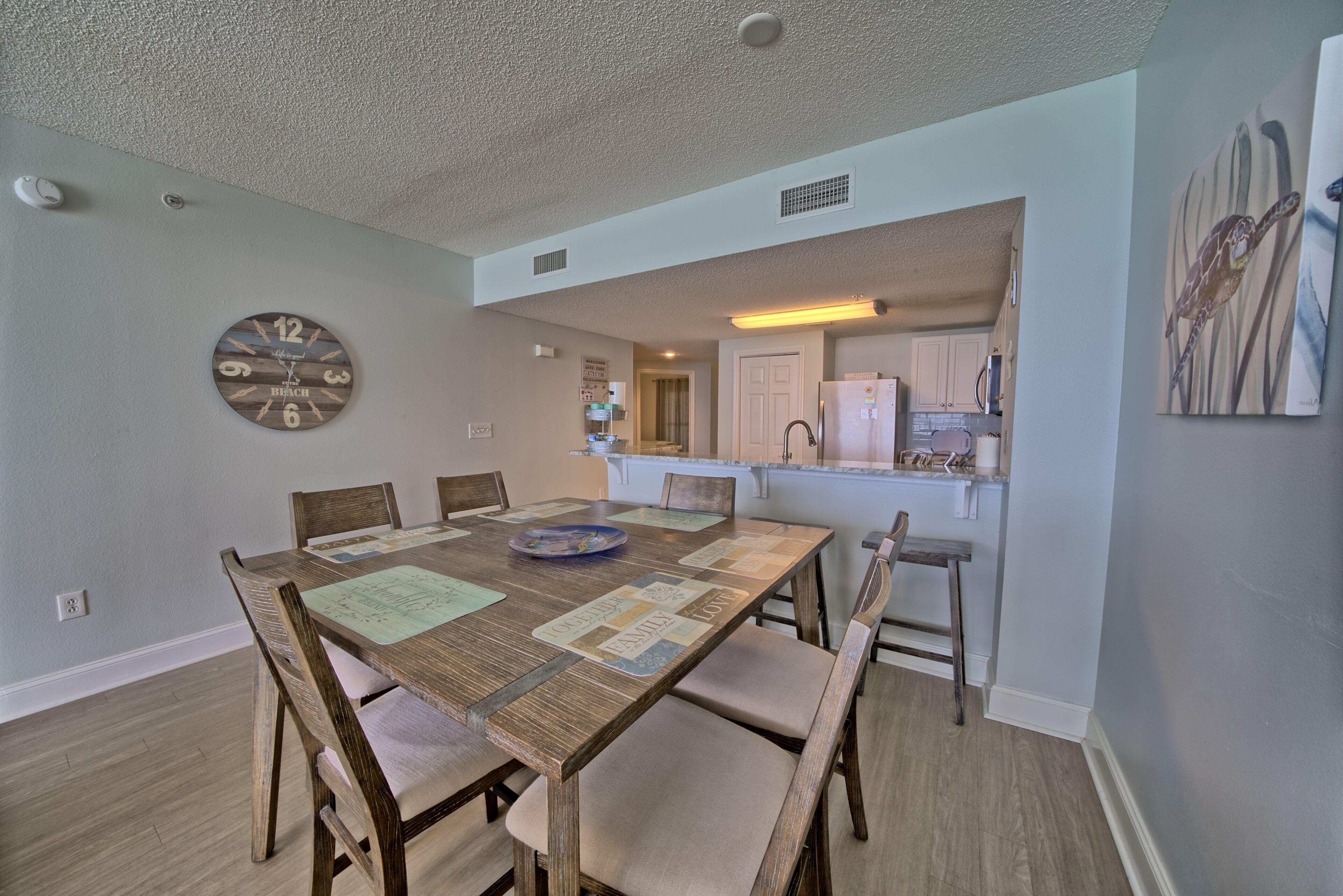 A dining area with a wooden table and chairs, a clock on the wall, and a kitchen in the background.