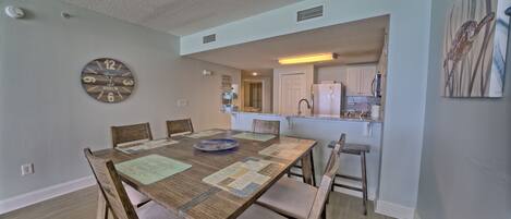 A dining area with a wooden table and chairs, a clock on the wall, and a kitchen in the background.