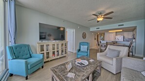 A spacious living room with a wooden coffee table, teal armchairs, and a dining area with wooden chairs and a kitchen in the background. - Play/Work right at the beach! Fast WiFi, Large balcony, Heated indoor pools (Panama City Beach)