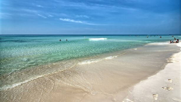 A beach with clear turquoise water, a sandy shore, and a pier in the distance. - Play/Work right at the beach! Fast WiFi, Large balcony, Heated indoor pools (Panama City Beach)