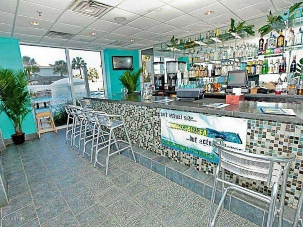 A bar area with a tiled backsplash, bar stools, and a counter with a computer and various bottles.