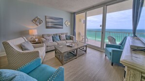 A spacious living room with a sofa, armchair, and a coffee table. A large window offers a view of the ocean. - Play/Work right at the beach! Fast WiFi, Large balcony, Heated indoor pools (Panama City Beach)