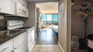 Une cuisine avec des meubles blancs, un four à micro-ondes et une cuisinière. Un couloir mène à un balcon d’où l’on a vue sur la mer.