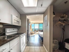 Une cuisine avec des meubles blancs, un four à micro-ondes et une cuisinière. Un couloir mène à un balcon d’où l’on a vue sur la mer.
