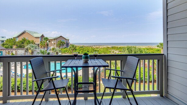 A balcony with a table and two chairs, overlooking a swimming pool and beachfront houses. - St Helena Island Condo - 1/2 Mi to Beach (Saint Helena Island)