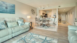 A modern living room with a light green sofa, a dining area with a white table and chairs, and a kitchen in the background. - St Helena Island Condo - 1/2 Mi to Beach (Saint Helena Island)