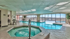An indoor swimming pool with a hot tub, surrounded by lounge chairs and a view of the city.
