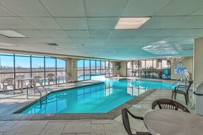 Une piscine intérieure offrant une vue dégagée sur les gratte-ciel de la ville, entourée de chaises longues et de tables.