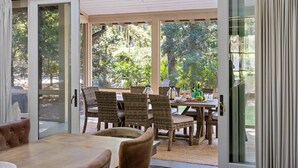 Een open woon-eetruimte met rieten stoelen en een houten tafel, met grote schuifdeuren naar een tuin.