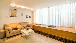 Ein modernes Wohnzimmer mit einem beigen Sofa, einem weißen Couchtisch und einem Holztische mit einem Buch. An der Wand hängen gerahmte Bilder und es gibt ein großes Fenster mit durchsichtigen Vorhängen.