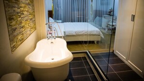 Ein modernes Badezimmer mit freistehender Badewanne, einer Duschkabine aus Glas und einem sichtbaren Schlafzimmer mit Bett und Vorhängen.